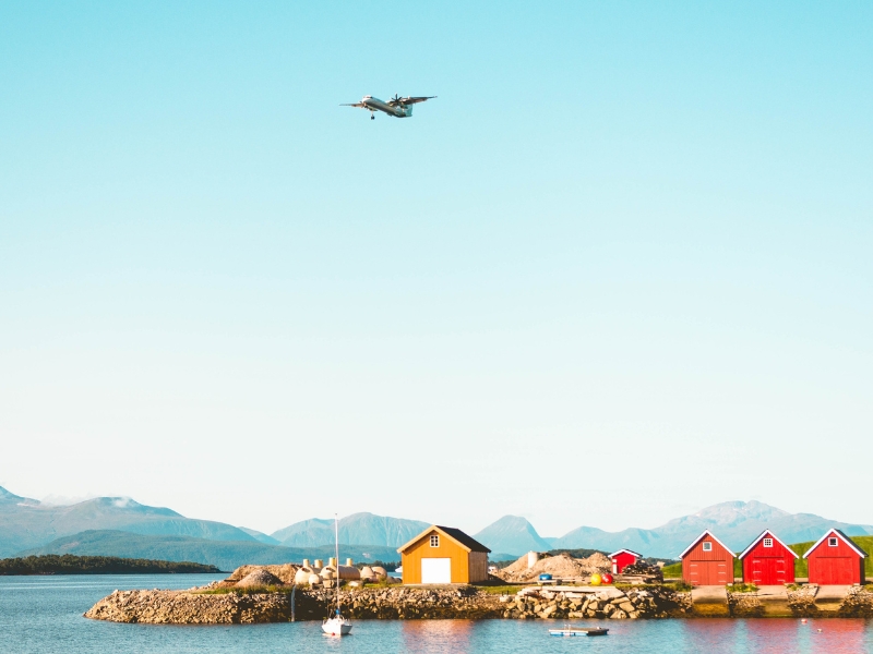 A small propeller aircraft aboue a fjord pier with small red and ywllow buildings and a white yacht.