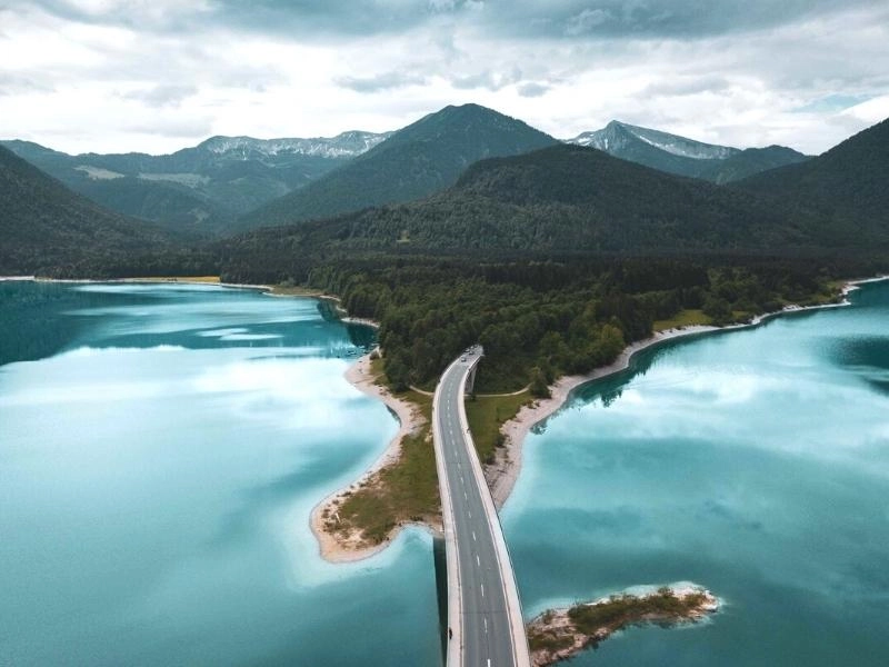 German road snaking across a blue lake with mountains in the background