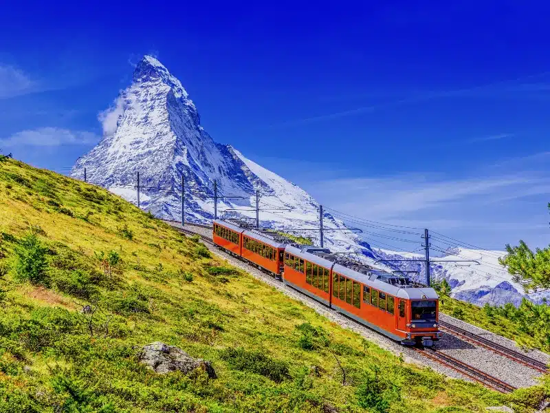 A red train passing in front of a large mountain through green fields